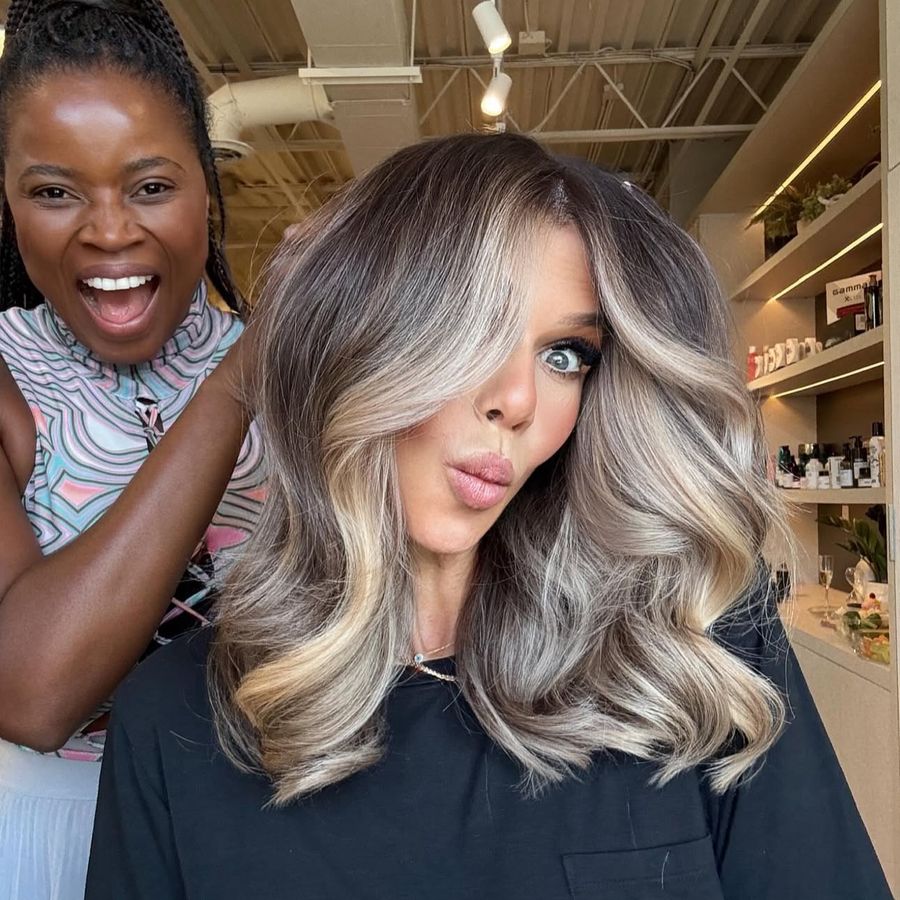A stylist laughing behind her client, who is showing a freshly finished blonde blowout in the salon.